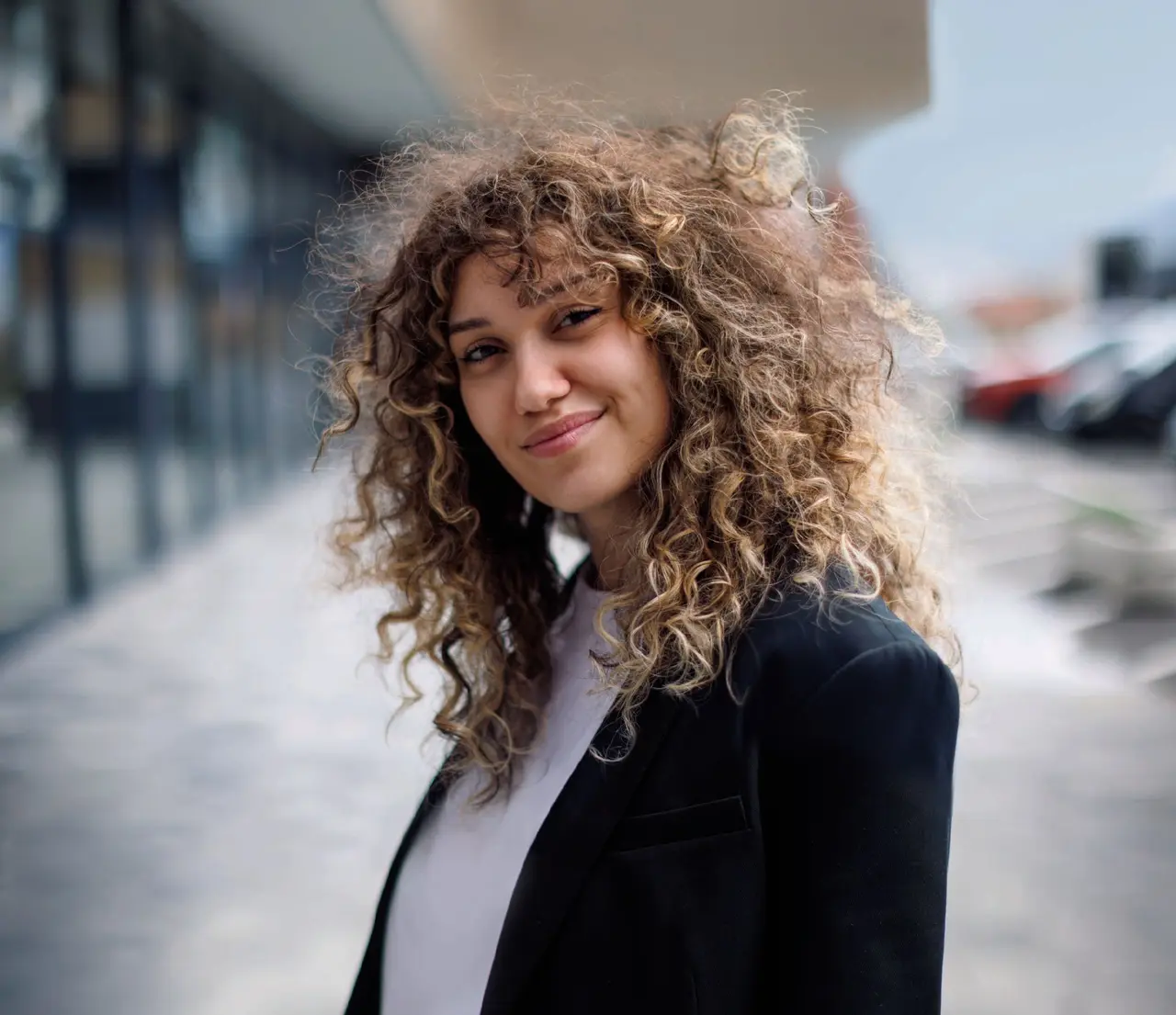 Une jeune professionnelle souriante aux cheveux bouclés pose en blazer noir.