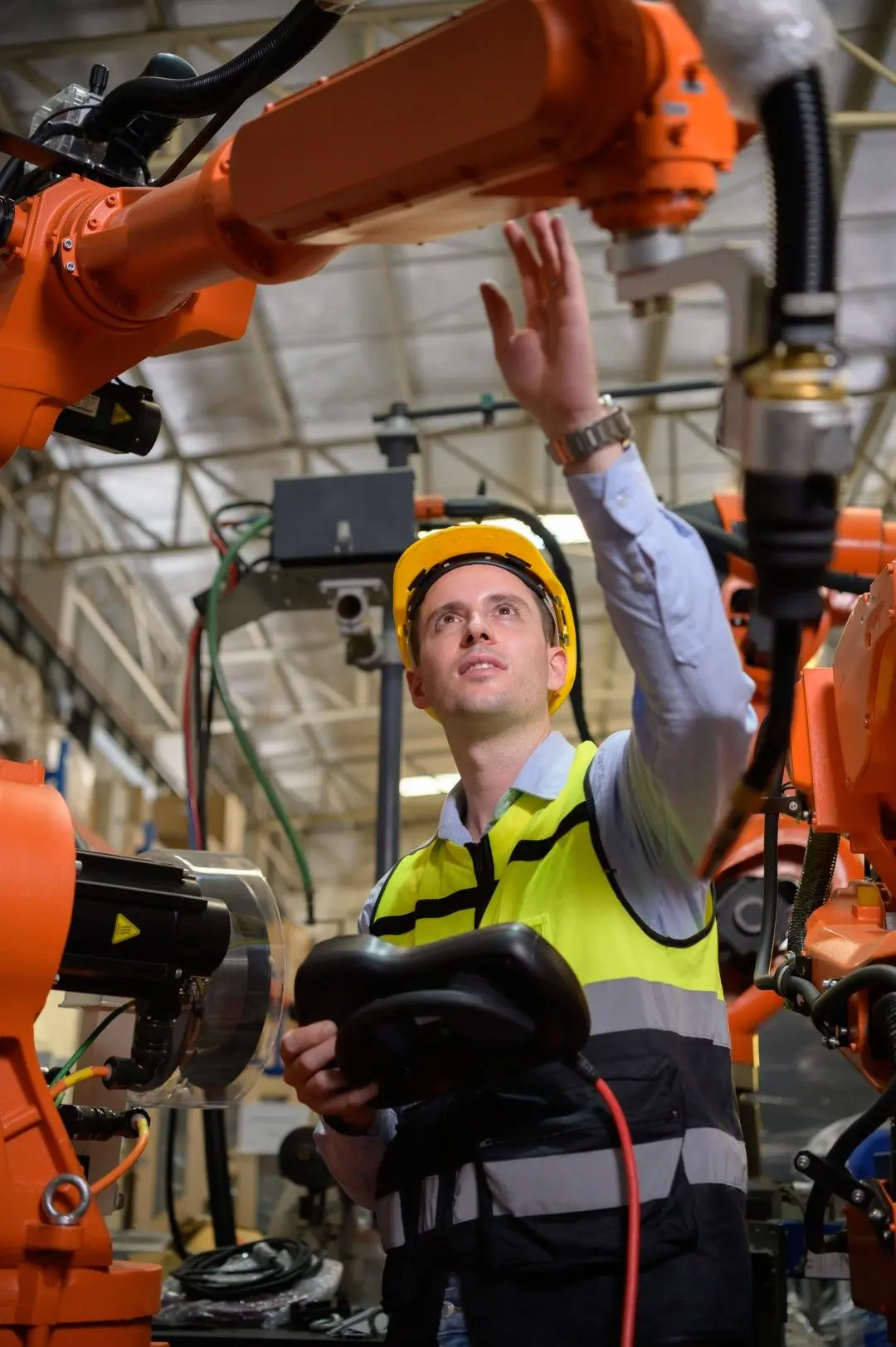 Un technicien industriel en casque de sécurité règle un bras robotisé orange dans un atelier de production.