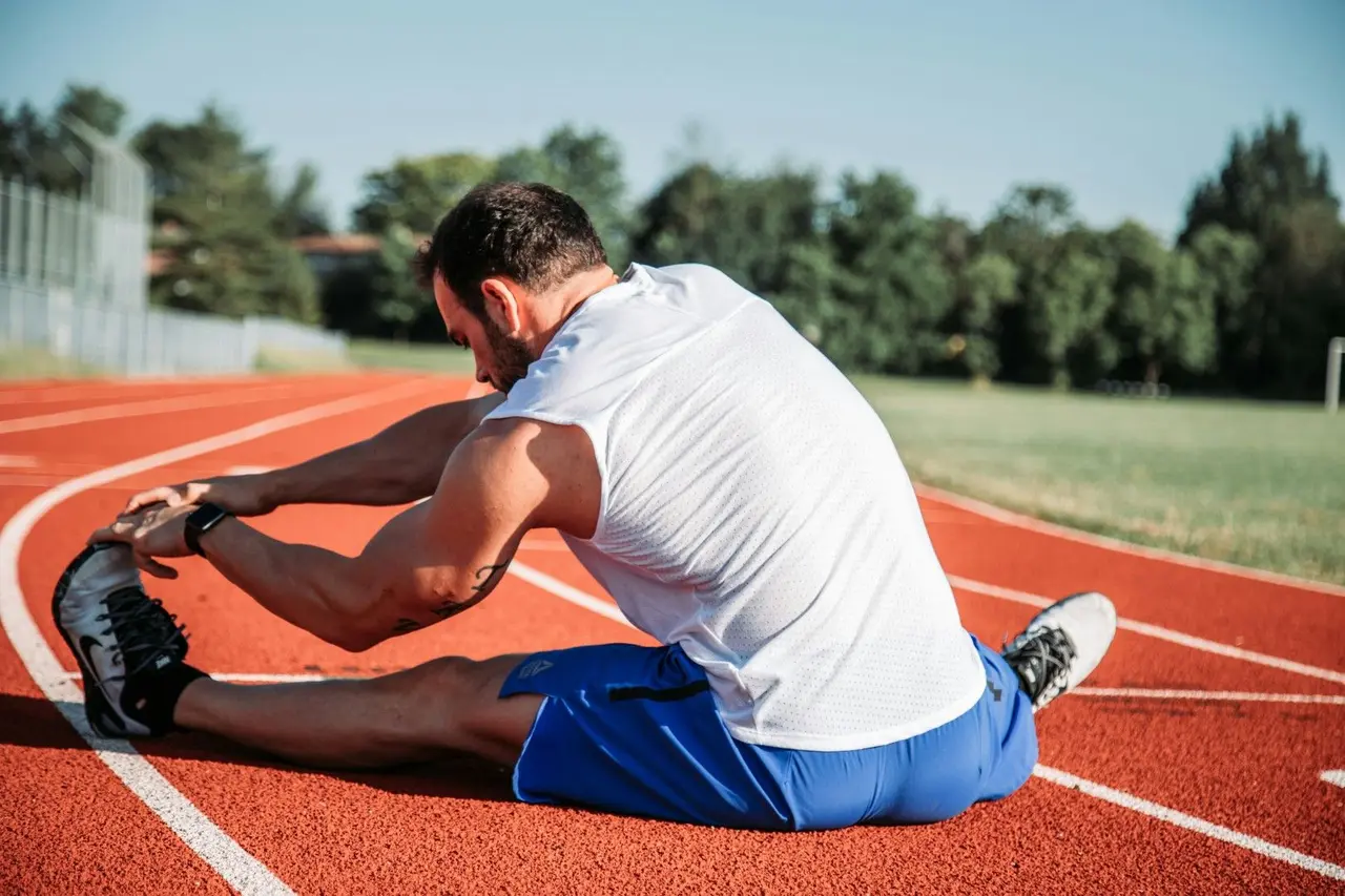 Un homme en tenue de sport s'étire au sol sur une piste d'athlétisme.