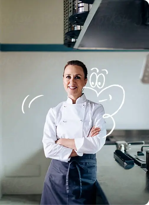 Une chef de cuisine souriante en veste blanche et tablier pose les bras croisés dans une cuisine professionnelle.