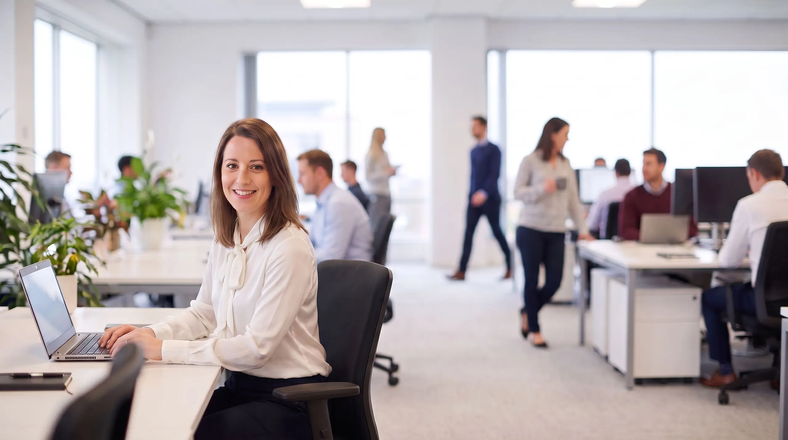 Une collaboratrice souriante travaille sur son ordinateur portable au sein d'un bureau en open space.