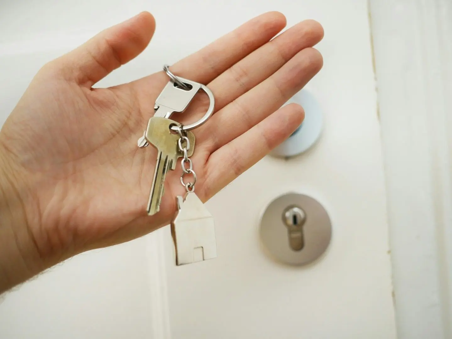 Une main tenant un trousseau de clés avec un porte-clé en forme de maison devant une porte blanche.