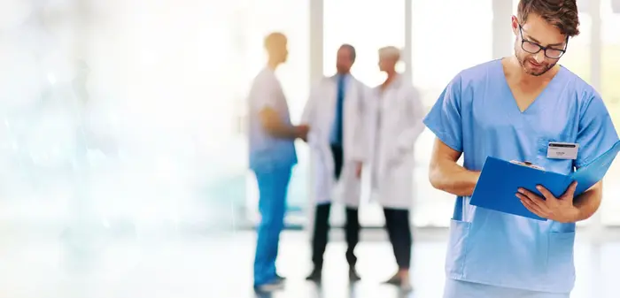 Un professionnel de santé en blouse bleue consulte un dossier, avec des collègues à l'arrière-plan.