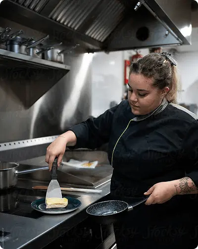 Une cuisinière en tenue professionnelle dresse une assiette avec une spatule dans une cuisine de restaurant.
