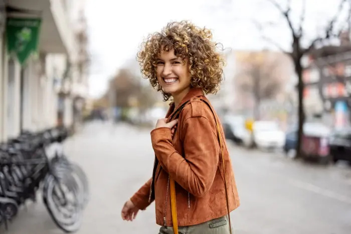 Une jeune femme souriante aux cheveux bouclés se retourne en marchant dans une rue.