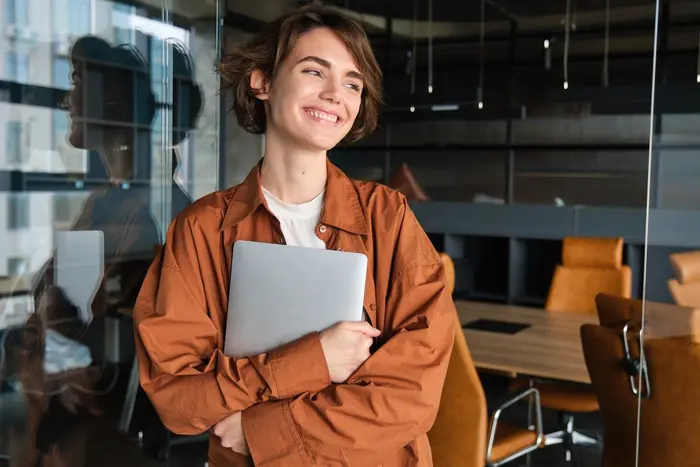 Une jeune femme souriante tient un ordinateur portable dans un bureau moderne.