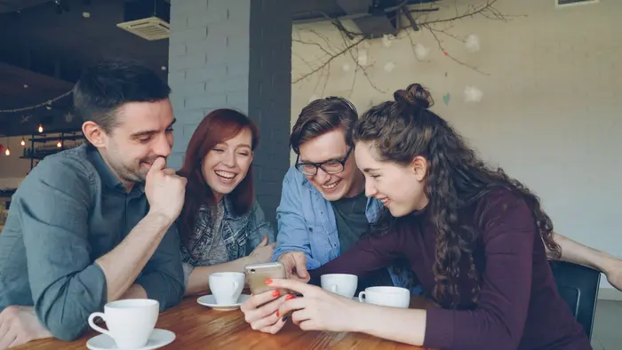 Quatre jeunes proches s’entraident et sourient autour d’un smartphone dans un café.