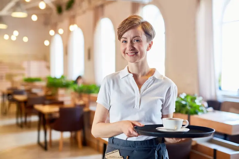 Une serveuse souriante en polo blanc tient un plateau avec une tasse de café dans un restaurant.